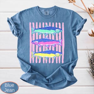 May include: Blue jean-colored t-shirt featuring a graphic of three colorful fish. The fish are in shades of green, pink, and yellow, set against a backdrop of pink vertical stripes. The shirt has a classic crew neck and short sleeves.