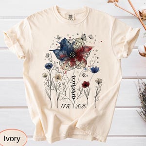 May include: Ivory-colored t-shirt featuring a floral design in red, white, and blue, with the word "america" in script. The design includes butterflies, stars, and the dates 1776 and 2026. The shirt is a comfortable, casual style.