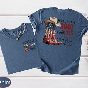 May include: Two denim-colored t-shirts. One features a small graphic of a cowboy boot and hat with the text "BROUGHT TO YOU COURTESY OF WHITE & BLUE". The other shirt displays a cowboy boot with an American flag design and the text "WE'LL PUT A BOOT IN YOUR ASS The American Way."