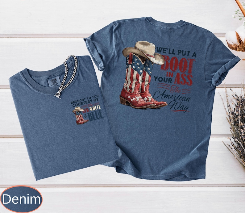 May include: Two denim-colored t-shirts. One features a small graphic of a cowboy boot and hat with the text "BROUGHT TO YOU COURTESY OF WHITE & BLUE". The other shirt displays a cowboy boot with an American flag design and the text "WE'LL PUT A BOOT IN YOUR ASS The American Way."