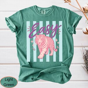 May include: Light green t-shirt with a pink tiger graphic and the word "Easy" in a cursive font. The tiger is walking forward, and the background has light blue vertical stripes.