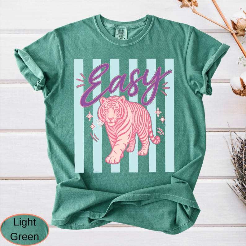May include: Light green t-shirt with a pink tiger graphic and the word "Easy" in a cursive font. The tiger is walking forward, and the background has light blue vertical stripes.