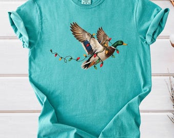 May include: A seafoam green short-sleeve t-shirt featuring a graphic of a mallard duck in flight, tangled in a string of colorful Christmas lights. The shirt has a crew neckline and a Comfort Colors brand label at the inner neck area.