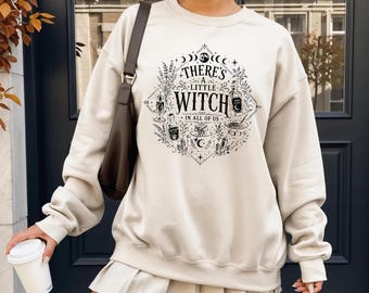 May include: A beige crewneck sweatshirt featuring a black graphic print that reads "THERE'S A LITTLE WITCH IN ALL OF US" surrounded by illustrations of moon phases, crystals, candles, a potion bottle, and a cauldron within a diamond-shaped frame.