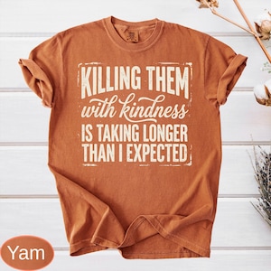 Op de afbeelding: Een roestoranje t-shirt met korte mouwen en witte tekst in een verweerde stijl die luidt: KILLING THEM with kindness IS TAKING LONGER THAN I EXPECTED. Het shirt heeft een ronde hals en een relaxte pasvorm met een zichtbaar merklabel in de kraag.