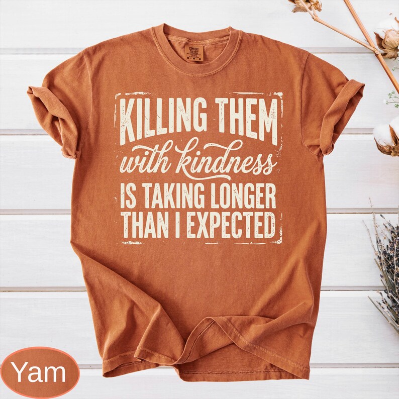 Op de afbeelding: Een roestoranje t-shirt met korte mouwen en witte tekst in een verweerde stijl die luidt: KILLING THEM with kindness IS TAKING LONGER THAN I EXPECTED. Het shirt heeft een ronde hals en een relaxte pasvorm met een zichtbaar merklabel in de kraag.