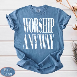 May include: Blue jean-colored t-shirt with the words "WORSHIP ANYWAY" in large, distressed white letters. The shirt is a crew neck style and is folded. The background is a white wooden surface.