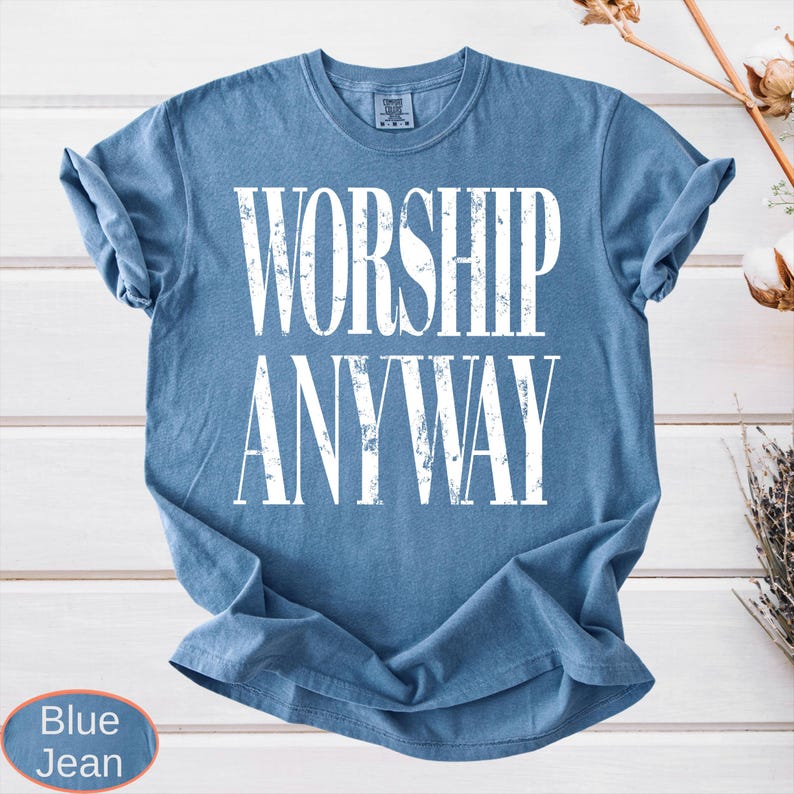 May include: Blue jean-colored t-shirt with the words "WORSHIP ANYWAY" in large, distressed white letters. The shirt is a crew neck style and is folded. The background is a white wooden surface.