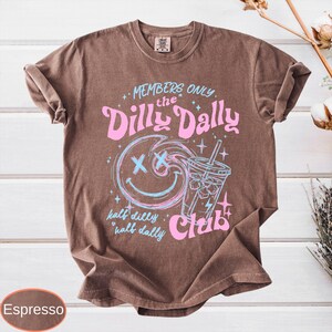 May include: Espresso-colored t-shirt with a graphic design. The design features the text "MEMBERS ONLY the Dilly Dally Club" in pink, along with a smiling face and a drink. The words "half dilly, half dally" are also included.