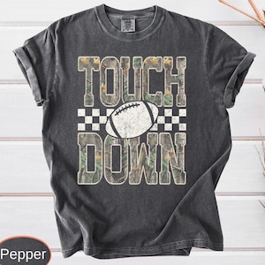 May include: A charcoal gray short-sleeve t-shirt featuring the text TOUCH DOWN in large block letters filled with a camouflage pattern. A white football graphic with black laces is centered between the words, set against a checkered background.