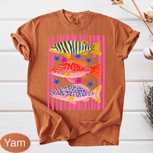 May include: A burnt orange t-shirt featuring a colorful graphic of three fish. The fish are in shades of green, red, and pink, with blue stars and pink star accents. The fish are set against a pink striped background.