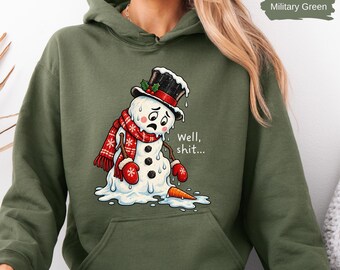 May include: A military green hooded sweatshirt featuring a graphic of a melting snowman wearing a black top hat, a red patterned scarf, and red mittens. The snowman has a sad expression and the text Well, shit... is printed next to it.