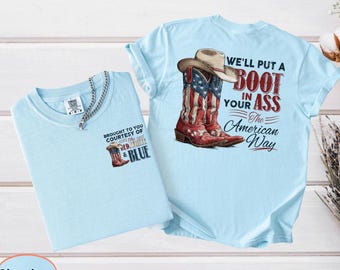May include: Two light blue t-shirts. One has a graphic of a cowboy boot with the American flag design and a cowboy hat, with the text "WE'LL PUT A BOOT IN YOUR ASS The American Way." The other shirt has a smaller boot graphic and the text "BROUGHT TO YOU COURTESY OF THE RED, WHITE & BLUE."