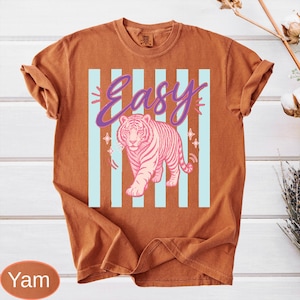 May include: A burnt orange t-shirt featuring a pink tiger graphic with the word "Easy" in purple script. The design includes light blue vertical stripes and star accents, creating a retro aesthetic.