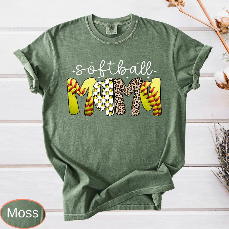 May include: Moss green t-shirt with the words "softball mama" in white script. The word "mama" is designed with a softball, leopard print, and softball stitching. The shirt is made of a soft material and has a relaxed fit.
