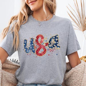 May include: A gray t-shirt with the word "USA" in a patriotic design. The letters are decorated with stars, floral patterns, and red and blue accents, with a watercolor effect.