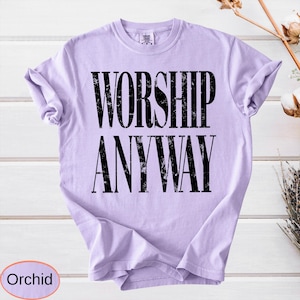 May include: A light purple t-shirt with the words "WORSHIP ANYWAY" printed in large, distressed black letters. The shirt is a solid color and has a crew neck. The word "Orchid" is in a circle at the bottom.