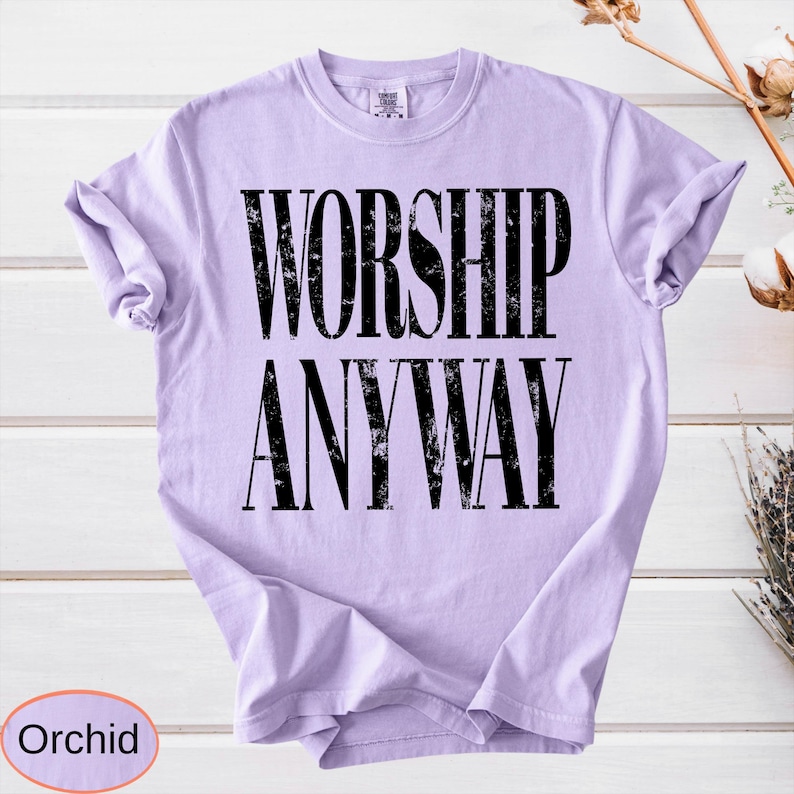 May include: A light purple t-shirt with the words "WORSHIP ANYWAY" printed in large, distressed black letters. The shirt is a solid color and has a crew neck. The word "Orchid" is in a circle at the bottom.