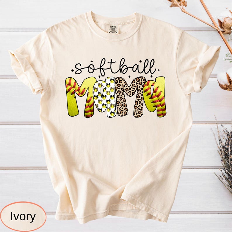May include: Ivory-colored t-shirt with the words "softball mama" in a playful design. The word "mama" features a mix of softball and leopard print patterns. The shirt is a comfortable, casual style.