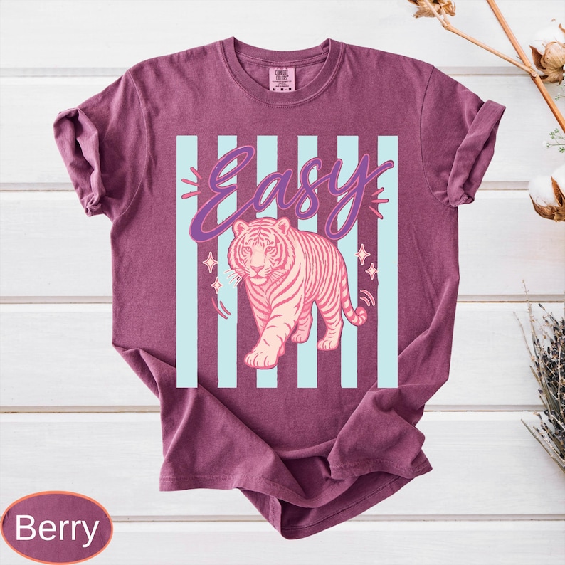 May include: Berry-colored t-shirt featuring a pink tiger illustration with the word "Easy" in a cursive font. The design includes light blue vertical stripes and star accents. The shirt has short sleeves.