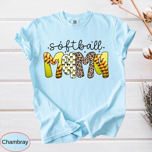 May include: Light blue t-shirt with the words "softball mama" in a playful design. The word "softball" is in black script, and "mama" is in yellow, leopard print, and softball-themed patterns. The shirt is made of a soft material.