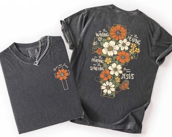 May include: Two charcoal gray t-shirts with floral cross designs. One shirt has a small cross with a flower and the text "Know me Jesus". The other shirt has a large floral cross design with the text "In the waiting, in the healing, in the hurting, in the searching, there was Jesus."