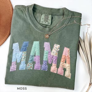 May include: A moss green t-shirt with the word "MAMA" appliqued in patchwork fabric. The patchwork letters feature various patterns and colors, including blue gingham, floral prints, and pink gingham. A silver necklace with a pendant rests on the shirt.