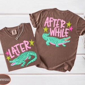 Puede incluir: Dos camisetas color espresso con dise&ntilde;os gr&aacute;ficos. Una camiseta muestra "LATER" con un caim&aacute;n verde azulado y estrellas, la otra dice "AFTER WHILE" con un caim&aacute;n y estrellas similares. Una cadena de plata est&aacute; en una camiseta.