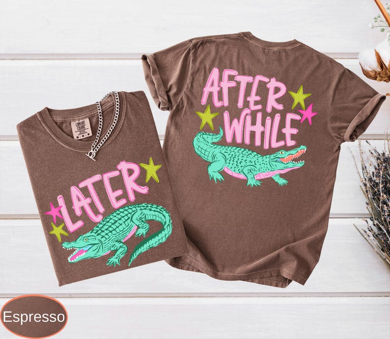 Puede incluir: Dos camisetas color espresso con dise&ntilde;os gr&aacute;ficos. Una camiseta muestra "LATER" con un caim&aacute;n verde azulado y estrellas, la otra dice "AFTER WHILE" con un caim&aacute;n y estrellas similares. Una cadena de plata est&aacute; en una camiseta.
