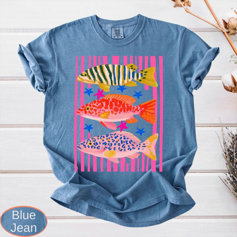 May include: Blue jean colored t-shirt featuring a colorful graphic of three fish. The fish are in shades of yellow, orange, and pink, with pink vertical stripes and blue stars. The shirt has a crew neck and short sleeves.
