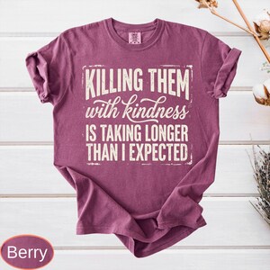 Op de afbeelding: Een bessenkleurig t-shirt met korte mouwen en de witte tekst "KILLING THEM with kindness IS TAKING LONGER THAN I EXPECTED" in een combinatie van blokletters en cursief schrift op de voorzijde. Het shirt heeft een ronde hals en een casual pasvorm.
