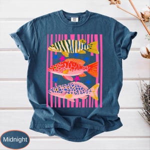 May include: Midnight blue t-shirt featuring a colorful graphic of three fish. The fish are set against a pink striped background with blue and pink star accents. The fish are yellow, orange, and pink with various patterns.