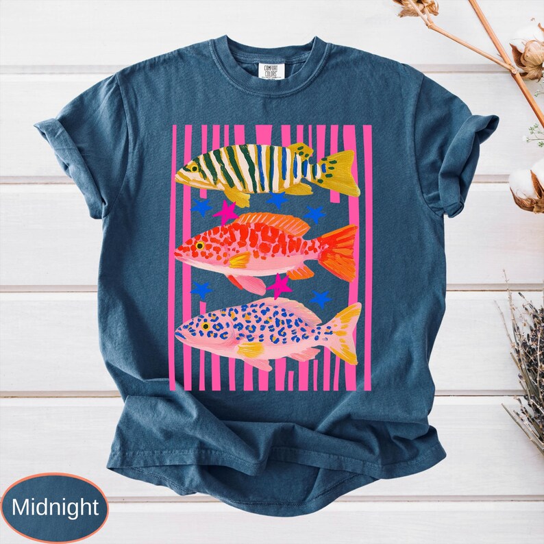 May include: Midnight blue t-shirt featuring a colorful graphic of three fish. The fish are set against a pink striped background with blue and pink star accents. The fish are yellow, orange, and pink with various patterns.