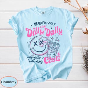 May include: Light blue t-shirt with the text "MEMBERS ONLY the Dilly Dally Club" in pink. The design includes a smiley face with an X for eyes, a drink, and the words "half dilly half dally".