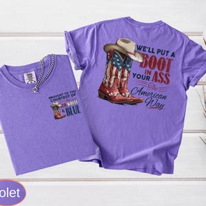 May include: Two violet t-shirts. One shirt has a small graphic of a cowboy boot with the text "BROUGHT TO YOU COURTESY OF RED WHITE & BLUE". The other shirt has a graphic of a cowboy boot with the American flag and the text "WE'LL PUT A BOOT IN YOUR ASS The American Way".