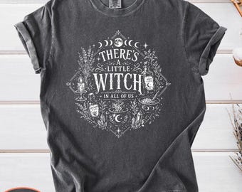 May include: A charcoal gray short-sleeve t-shirt featuring a white graphic print of a diamond-shaped frame containing botanical elements, a candle, potion bottles, a cauldron, and the text "THERE'S A LITTLE WITCH IN ALL OF US" in a stylized font.