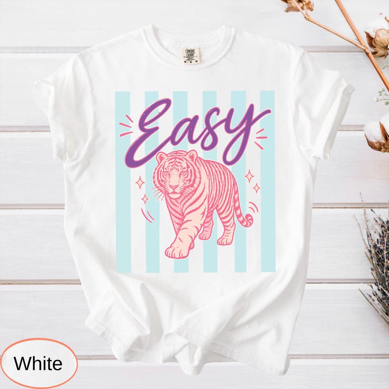 May include: White t-shirt with a pink tiger graphic and the word "Easy" in purple script. The tiger is walking forward against a backdrop of light blue vertical stripes. The shirt has a classic crew neck and short sleeves.