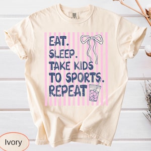 May include: Ivory-colored t-shirt with the text "EAT. SLEEP. TAKE KIDS TO SPORTS. REPEAT" in dark blue. The design includes a pink and white striped background, a bow, and a drink with ice. The shirt is a Comfort Colors brand.