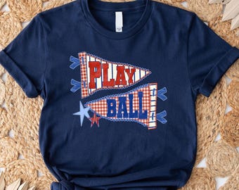May include: Navy blue t-shirt with a patriotic design. The shirt features two pennant-shaped graphics with the words "PLAY" and "BALL" in red and blue, with star and gingham patterns. A casual tee for sports fans.