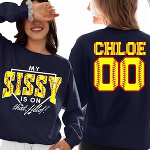 May include: Navy blue sweatshirt with softball-themed text and graphics. The front displays "MY SISSY IS ON that field!" in yellow and white. The back features "CHLOE" and the number "00" in a similar softball design. A sporty apparel item.