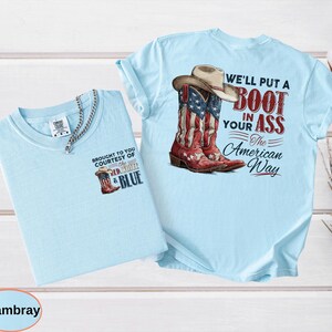 May include: Two light blue t-shirts. One features a graphic of a cowboy boot with the American flag design and the text "BROUGHT TO YOU COURTESY OF THE RED, WHITE & BLUE". The other shirt has a similar graphic of a cowboy boot and a hat with the text "WE'LL PUT A BOOT IN YOUR ASS The American Way".