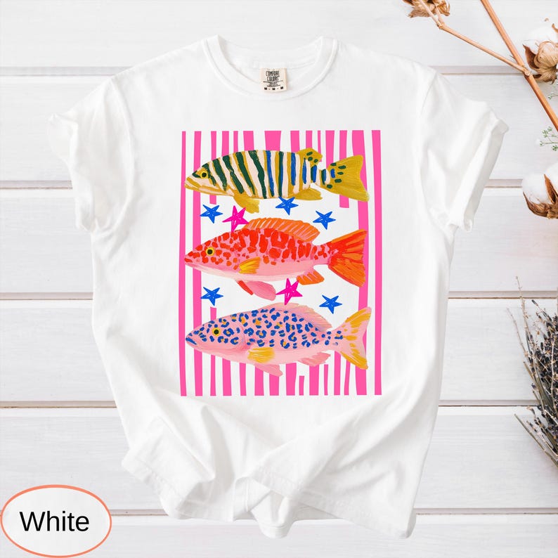 May include: White t-shirt featuring a colorful fish design. The graphic includes three fish in yellow, red, and blue patterns, set against pink stripes and blue and pink stars. A casual, comfortable tee.
