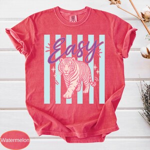 May include: Watermelon-colored t-shirt with a tiger graphic. The design features a pink tiger walking in front of light blue vertical stripes. The word "Easy" is written in a cursive font above the tiger.
