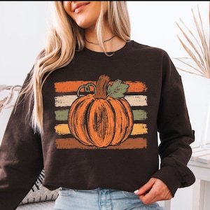 May include: A dark brown long-sleeve sweatshirt featuring a central graphic of an orange pumpkin with a green leaf and stem. The pumpkin is layered over a background of horizontal, distressed-texture stripes in shades of orange, cream, and green.