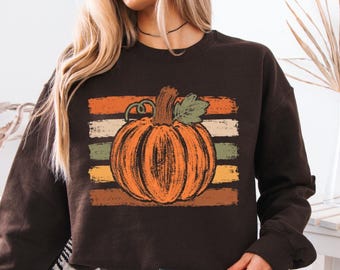 May include: A dark brown long-sleeve sweatshirt featuring a central graphic of an orange pumpkin with a green leaf and stem. The pumpkin is layered over a background of horizontal, distressed-texture stripes in shades of orange, cream, and green.