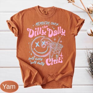May include: A burnt orange t-shirt with rolled sleeves. The front features a graphic with the text "MEMBERS ONLY the Dilly Dally Club". The graphic includes a smiling face, a drink, and the words "half dilly half dally" in pink and blue.