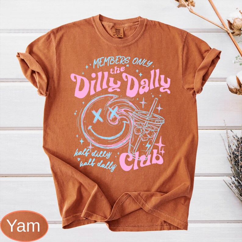 May include: A burnt orange t-shirt with rolled sleeves. The front features a graphic with the text "MEMBERS ONLY the Dilly Dally Club". The graphic includes a smiling face, a drink, and the words "half dilly half dally" in pink and blue.