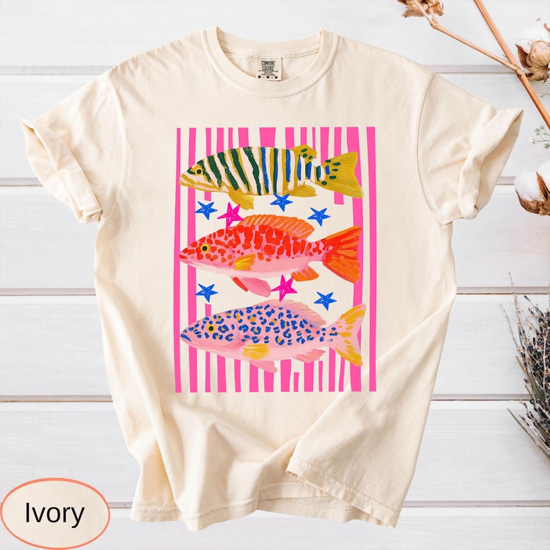 May include: Ivory-colored t-shirt featuring a colorful fish design. The graphic includes three fish in yellow, red, and blue, set against a pink striped background with blue and pink stars. The shirt has a classic crew neck.