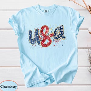 May include: Light blue t-shirt with the word "USA" in a patriotic design. The letters are decorated with stars, dots, and floral patterns in red, white, and blue, with gold outlines. The shirt is made of a soft material.