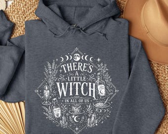 May include: A dark heather gray hooded sweatshirt featuring a white graphic print. The design includes the text "THERE'S A LITTLE WITCH IN ALL OF US" surrounded by illustrations of a candle, lavender, a potion bottle, a cauldron, and moon phases.